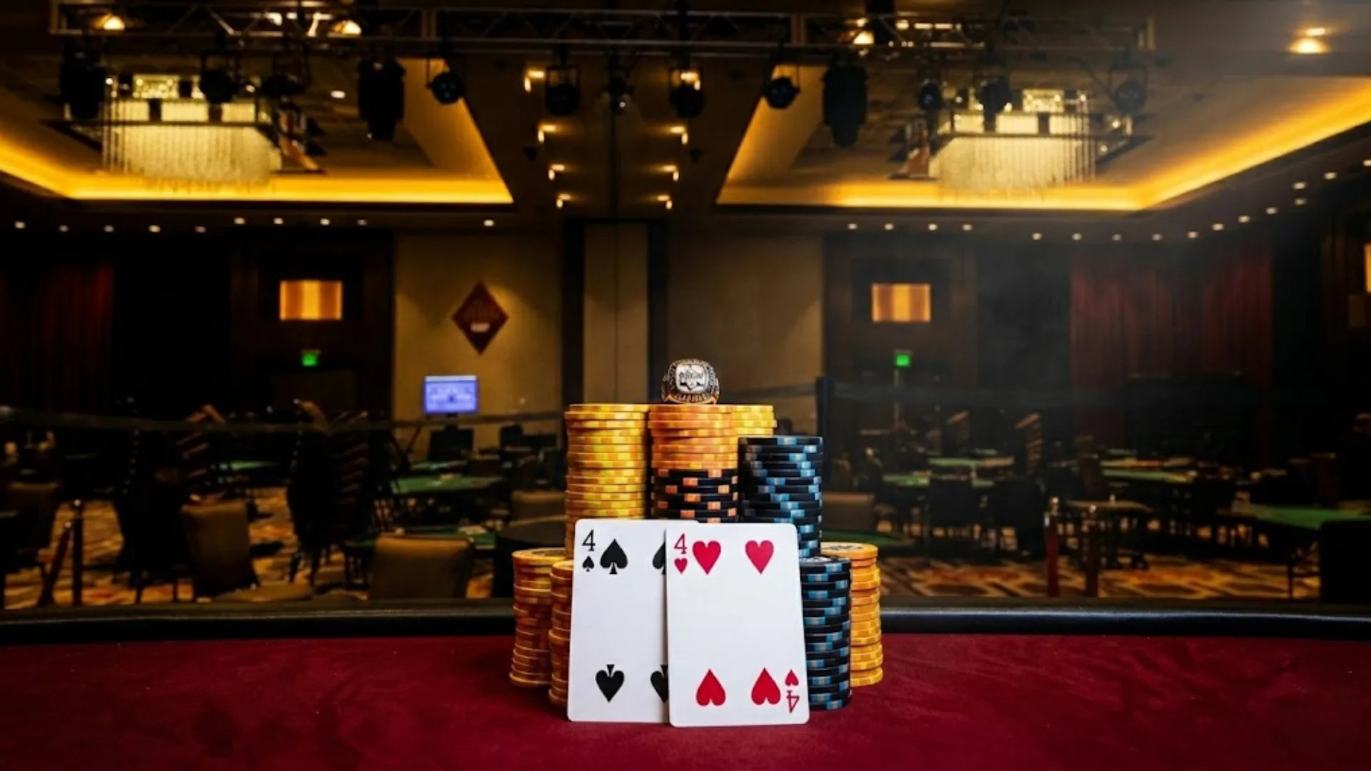 豪華なカジノのポーカー・トーナメント決勝テーブルのクローズアップ。高く積まれたイエローとブルーのチップ、スペードの4とハートの4の勝利ハンド、そしてその上に置かれた誰もが憧れる精巧なポーカー・チャンピオン・リング。